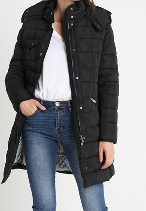 Femme portant un long manteau matelassé noir avec fermeture éclair et boutons-pression, chemise blanche et jean bleu, main dans la poche, debout devant un fond blanc.