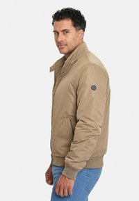 Chaqueta bomber color beige con cierre frontal, puños y dobladillo acanalados, bolsillos laterales y un parche de logo redondo en la manga izquierda. Lleva puesta con jeans azules.