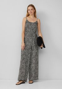 Schwarz-weiß gemusterter Jumpsuit mit lockerer Passform, dünnen Trägern und gebundenem Taillengürtel. Das Model hält eine strukturierte schwarze Handtasche.
