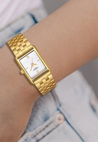 Zelta tonēta taisnstūra pulksteņi ar baltu teksturētu ciparnīcu, zelta rokturiem un aušanas metāla siksnu. Vienkārša aizdare.