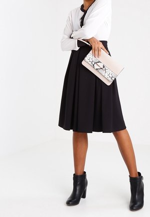 Gonna plissettata nera abbinata a una blusa bianca, indossando stivaletti neri alla caviglia e tenendo una pochette testurizzata con motivo a serpente.