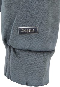 Poignet de manche en tissu gris avec une petite étiquette métallique rectangulaire embossée du mot « Dropside » attachée près de la couture.