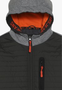 Chaqueta con capucha en negro y gris con forro naranja, textura acolchada, cierre frontal y un bolsillo en el pecho con cremallera y tirador naranja.