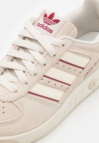 Beige suède sneakers met witte en bordeauxrode gestreepte accenten, met een gestructureerde zool, ronde veters en een logo op de tong.