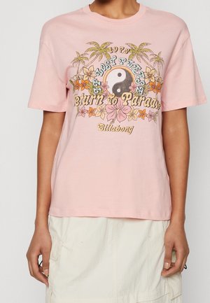 Persoon die een roze t-shirt draagt met een yin-yang, palmbomen, bloemen en de tekst "1973 Lost Please Return to Paradise Billabong" op de voorkant.
