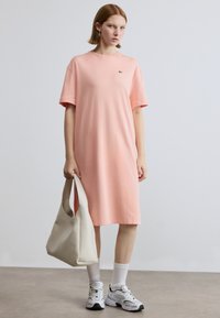 Φόρεμα από βαμβακερό ύφασμα σε ανοιχτό ροζ χρώμα, με κοντά μανίκια, στρογγυλή λαιμόκοψη και μικρό λογότυπο. Συνδυασμένο με μπεζ τσάντα και λευκά αθλητικά παπούτσια.