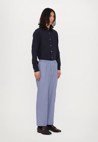 Marineblaues Button-Up-Hemd aus Leinen, hellblaue gefaltete Hose mit elastischem Bund, braune Slipper mit goldenen Akzenten. Einfarbiger weißer Hintergrund.