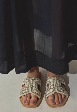 Picioare îmbrăcate în sandale albe și aurii cu mărgele, sub o fustă lungă neagră transparentă, pe un podea deschisă la culoare.