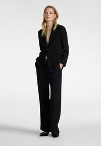 Donna in piedi con le mani nelle tasche, indossa un blazer nero oversize, pantaloni larghi, scarpe a punta nere, una collana d'oro e orecchini a forma di stella.