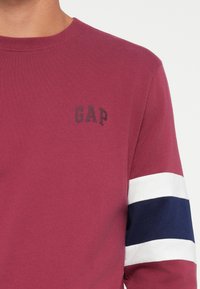 Maroon long-sleeve shirt with dark gray "GAP" logo. Features white and navy stripes on sleeves, crafted from soft cotton fabric.
