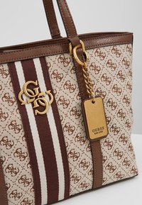 Sac cabas Guess beige et marron avec motif logo répété, garniture en cuir marron, emblème logo doré et étiquette dorée indiquant "GUESS EST 1981".