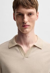 Polo shirt tricotat bej, cu un țesătură texturată, având un decolteu în V și o guler plat. Design ajustat, cu mâneci scurte.