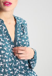 Tealblus med djurtryck i vitt, med en djup V-ringning och knapprad, långa ärmar och en slät tygtextur.