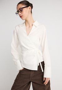 Λευκό πουκάμισο τύπου wrap με γιακά, μακριά μανίκια και λεπτομέρεια ζώνης στη μέση, συνδυασμένο με καφέ παντελόνι από κορντόρι. Λείο ύφασμα, ελαφρύ υλικό.