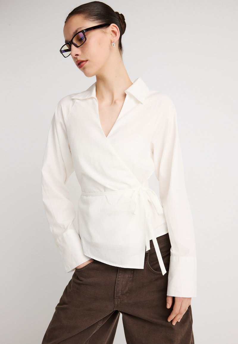 Λευκό πουκάμισο τύπου wrap με γιακά, μακριά μανίκια και λεπτομέρεια ζώνης στη μέση, συνδυασμένο με καφέ παντελόνι από κορντόρι. Λείο ύφασμα, ελαφρύ υλικό.