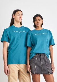 Türkiissinist puuvillased T-särgid, millel on "TOMMYJEANS" logo. Mugav lõige, ümar kaelus, lühikesed varrukad. Paremal olev mudel kannab musta teksaseelikut.