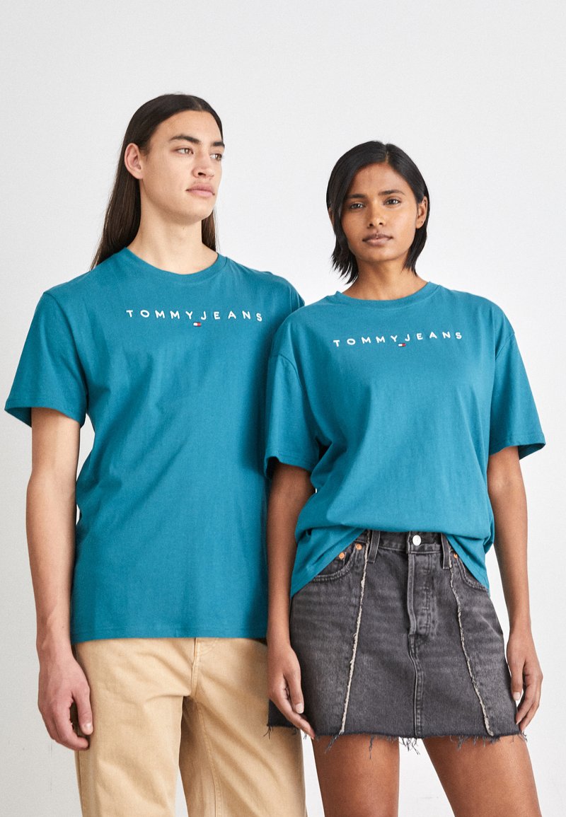Türkiissinist puuvillased T-särgid, millel on "TOMMYJEANS" logo. Mugav lõige, ümar kaelus, lühikesed varrukad. Paremal olev mudel kannab musta teksaseelikut.