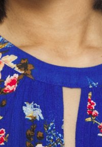 Modrá texturovaná tkanina s květinovými vzory v růžové, bílé a zelené barvě. Obsahuje výstřih ve tvaru klíčové dírky a nabrané detaily na ramenech.