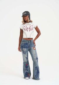 Lyhyttaskuinen valkoinen t-paita, jossa on punainen graafinen painatus, yhdistettynä sinisiin farkkuihin, joissa on brodeeratut lohikäärmeet ja kukkakuviot. Valkoiset lenkkarit viimeistelevät asun.