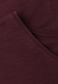 Gros plan sur un tissu bordeaux montrant une texture fine en maille et une couture précise le long d’un bord plié.