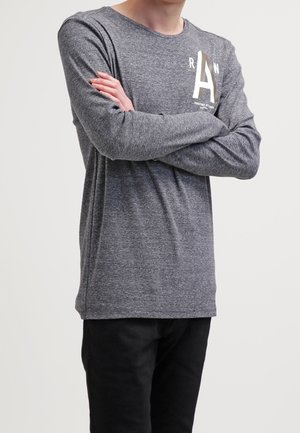 Langärmliges graues T-Shirt aus strukturiertem Stoff, mit einem weißen Druckbuchstaben "A" und kleinem Text auf der linken Seite, kombiniert mit schwarzen Hosen.