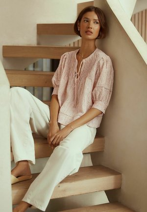 Junge Frau mit kurzen Haaren, die auf einer Holztreppe sitzt und ein lockeres, rosa besticktes Oberteil, weiße Hosen und beigen Schlupfschuhen trägt.