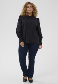Camisa negra de botones con rayas verticales texturizadas, mangas largas con puños fruncidos, combinada con jeans de mezclilla oscuros y bailarinas negras.