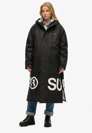 Zwarte lange jas met een capuchon, met grote witte logo's aan de onderkant, voorzakken en textiel met structuur. Gedragen over jeans en laarzen.