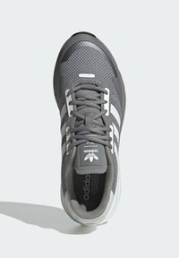 Šedá športová obuv s prednou časťou z technickej sieťoviny, biele pásiky a gumová podrážka. Obsahuje šnúrky a logo na jazyku a vonkajšej strane.