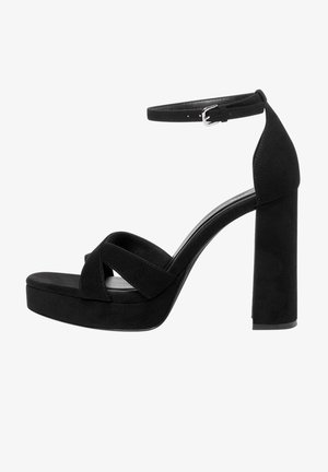 Sandalias de tacón alto de ante negro con plataforma, con tiras cruzadas y una correa en el tobillo con hebilla, sobre un tacón cuadrado grueso.