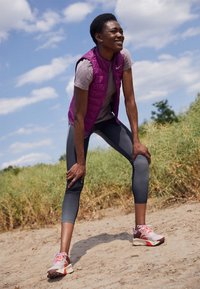 Gilet rembourré violet par-dessus un haut à manches courtes gris clair, associé à des leggings grisés ajustés et des chaussures de sport multicolores, dans un environnement extérieur.