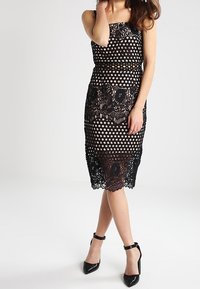 Musta olkaimeton mekko, jossa on pilkkuikkuna ja pitsiyksityiskohtia helmassa. Kiiltäviä mustia korkokenkiä täydentää asun.