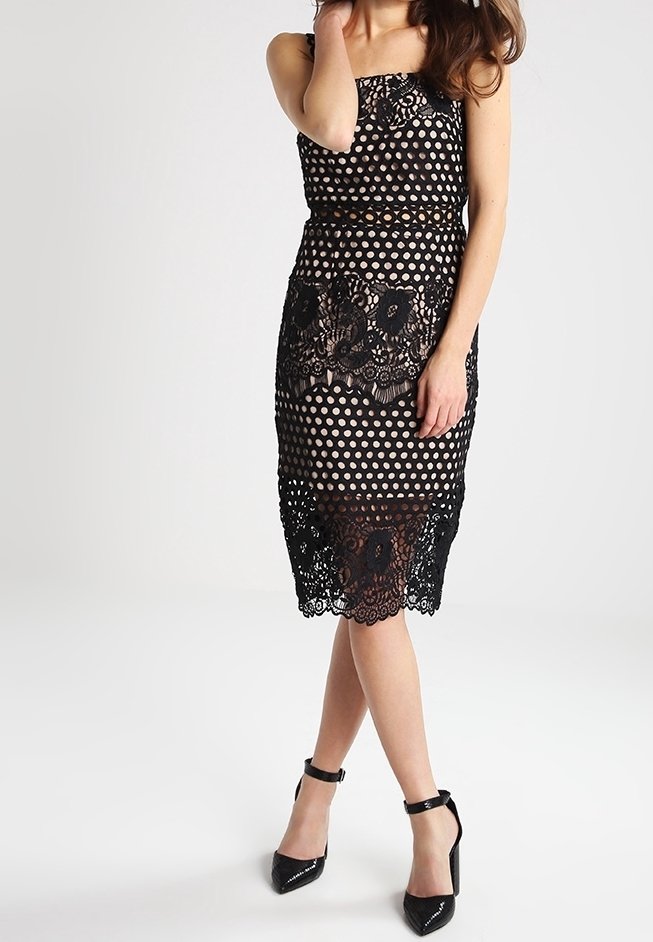 Černé šaty bez ramínek s puntíkovaným vzorem a krajkovým zdobením na okraji. Lesklé černé vysoké podpatky dokončují vzhled.