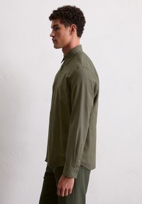 Man in olijfgroene lange mouwen blouse en bijpassende broek staat in profiel tegen een eenvoudige witte muur.