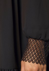 Blouse noire en tissu lisse avec des manches cloche en dentelle texturée, ornée d'un motif festonné aux poignets.