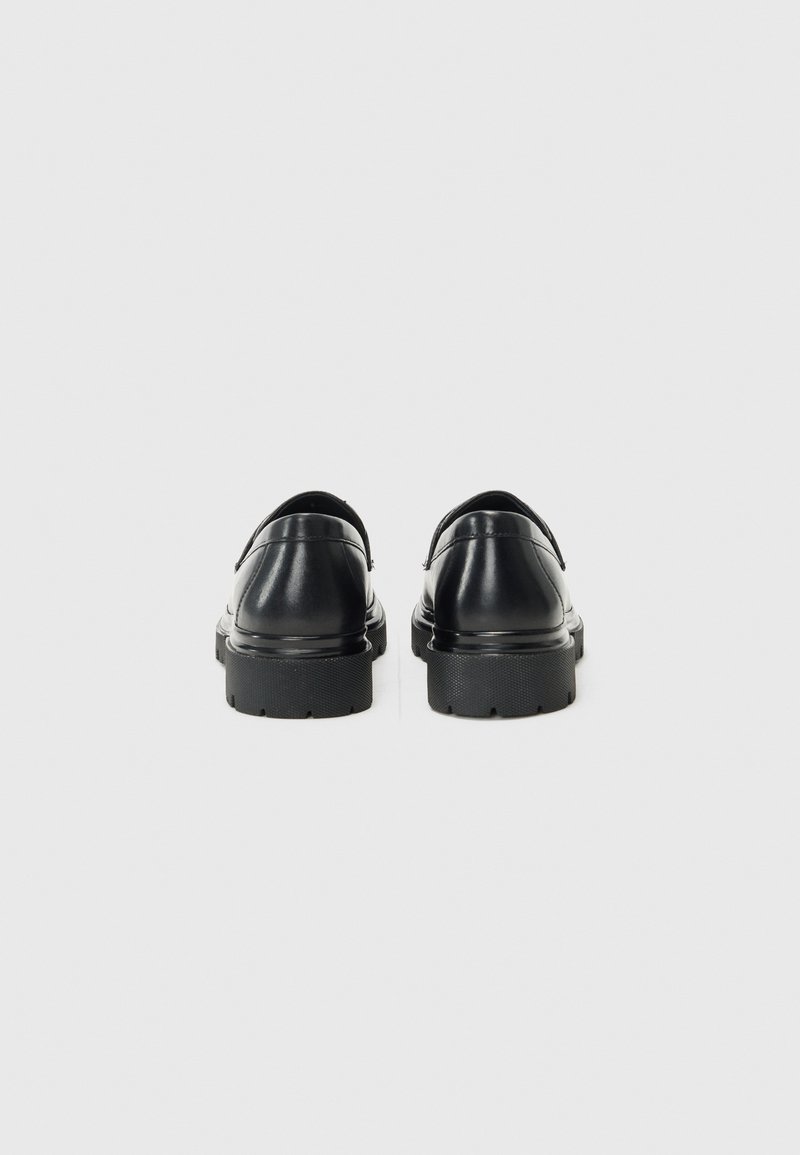 Schwarze Lederschuhe mit glatter Oberfläche, runder Form, flacher, dicke Sohle und minimalistischem Design. Leicht erhöhter Fersenbereich und strukturierte Außensohle.