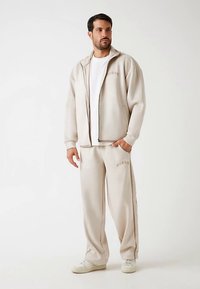 Beige Jacke zum Reißverschluss und passende Hose aus weichem Material; mit dezenten Logoakzenten und einem lockeren Schnitt.