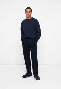 Sudadera de cuello redondo en azul marino combinada con pantalones de pana en azul marino oscuro. Modelo de pie con las manos en los bolsillos, usando calzado negro.
