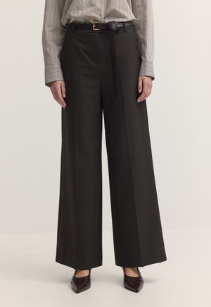Schwarze weite Hosen mit schwarzem Gürtel, getragen zu einem beige gestreiften Hemd und spitzen braunen Schuhen, die unten sichtbar sind.