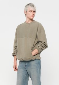 Fleece sweatshirt i olivengrønn, med mannekjøring, droppede skuldre, ribbestrikkede mansjetter og en horisontal søm foran. Brukt sammen med lyseblå jeans.