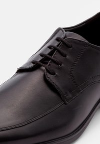 Chaussure habillée en cuir noir avec une finition lisse, bout rond, design à lacets à six œillets et détails de couture subtils le long des côtés.