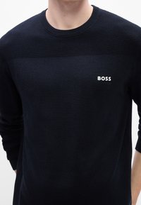 Homme portant un pull en maille noir avec une texture sur la partie supérieure de la poitrine et le logo "BOSS" en blanc sur le côté gauche de la poitrine.