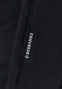 Bolsa de tela negra con superficie texturizada y una etiqueta blanca con el logo "CONVERSE" adjunta a un lado. Diseño simple y forma funcional.