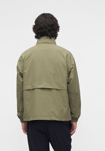 Personne aux cheveux bruns ondulés portant une veste kaki avec une fente dans le dos, debout devant un fond gris clair uni.