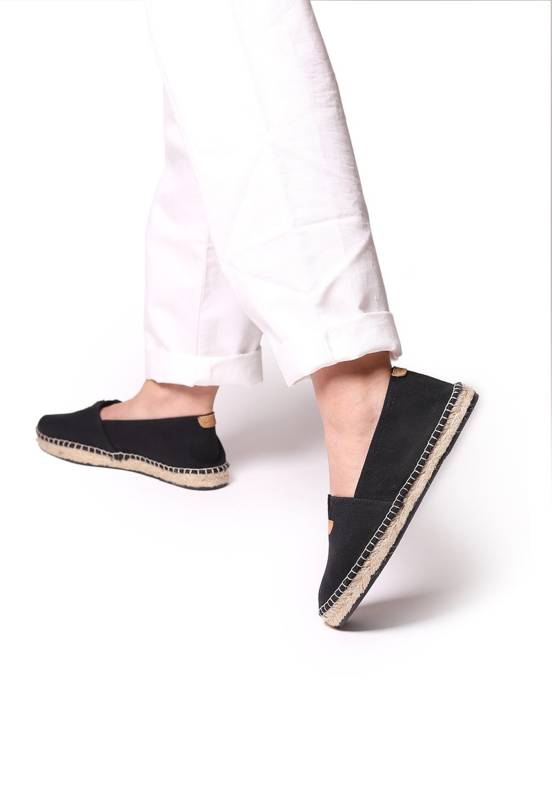 Černé plátěné espadrilky s jutovou podrážkou, s plochým designem a prošívaným okrajem. Na zadní straně se nachází malá béžová akcentová nálepka.