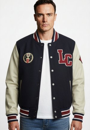Uomo che indossa una giacca varsity blu navy e crema con polsini a righe rosse e bianche e toppe con le lettere "LC" e un emblema alato.
