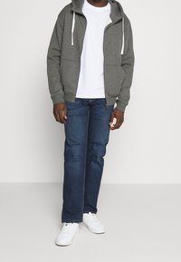 Grauer Reißverschluss-Hoodie mit weißen Kordeln, getragen über einem weißen T-Shirt, kombiniert mit dunkelblauen Jeans und weißen Turnschuhen. Einfaches und lässiges Design.