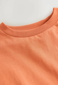 Koszulka z krótkim rękawem typu crewneck w stonowanym pomarańczowym kolorze, z gładką teksturą i ribbowanym dekoltem, ułożona na jasnym tle.
