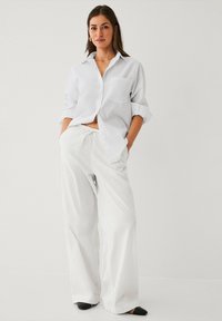 Camicia bianca in lino con maniche arrotolate, abbinata a pantaloni larghi bianchi in lino. Scarpe chiuse nere, design semplice.