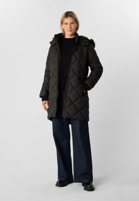 Cappotto imbottito nero a trapunte con cappuccio e bordo in pelliccia sintetica, dotato di zip frontale e tasche laterali, indossato sopra un maglione nero e jeans scuri.